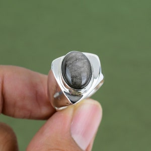 Puede incluir: Un anillo de plata con una gran piedra preciosa negra de forma ovalada. El anillo tiene un diseño único con una banda curva y un engaste elevado para la piedra.