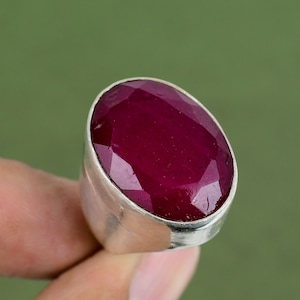 Peut inclure: Une bague en argent avec une grande pierre précieuse en rubis facetté de forme ovale. Le rubis est de couleur rouge foncé et a un haut poli.
