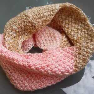 May include: A handmade infinity scarf in a two-tone design. The top half is a light tan color, and the bottom half is a soft pink. The scarf is made of a textured, knitted material, perfect for cooler weather.