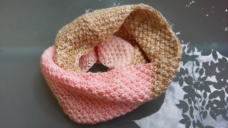 May include: A handmade infinity scarf in a two-tone design. The top half is a light tan color, and the bottom half is a soft pink. The scarf is made of a textured, knitted material, perfect for cooler weather.