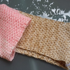 May include: Two handmade crochet scarves. One is folded and pink, the other is tan and unfolded. Both have a textured, diagonal stitch pattern. The scarves are displayed on a reflective surface.
