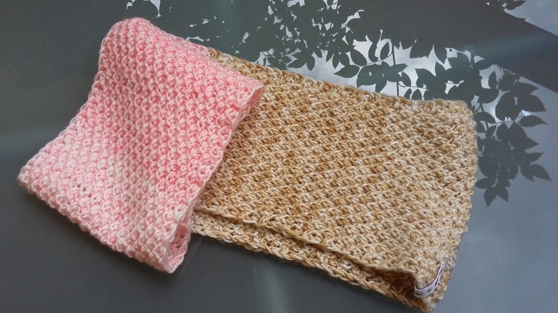 May include: Two handmade crochet scarves. One is folded and pink, the other is tan and unfolded. Both have a textured, diagonal stitch pattern. The scarves are displayed on a reflective surface.