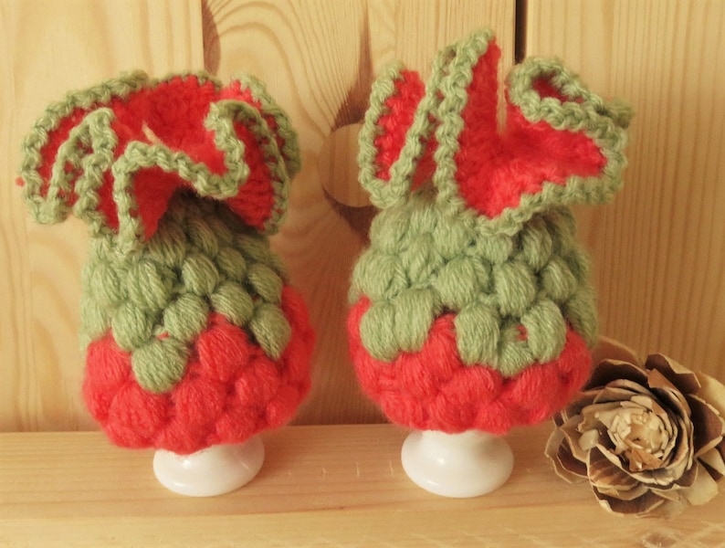 May include: Two handmade, crocheted egg cozies. Each cozy features a red base, a green textured body, and a ruffled red and green top. The cozies sit on white egg cups, perfect for adding a touch of whimsy to your breakfast table.