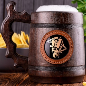 Puede incluir: Una jarra de cerveza grande de madera oscura llena de cerveza espumosa. La jarra tiene un asa decorativa y un parche de cuero con un diseño dorado. Un cuenco de patatas fritas está en el fondo.