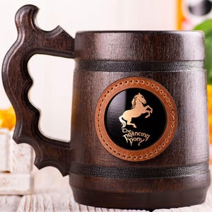 Op de afbeelding: Een donkerbruine houten bierkan met een leren patch met een gouden silhouet van een paard en de tekst "The Prancing Pony".