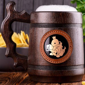 May include: A brown wooden beer stein with a leather patch featuring a cartoon creature wearing a crown and holding a beer mug. The stein is filled with a white foamy liquid.
