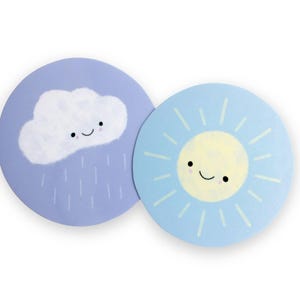 Puede incluir: Dos posavasos redondos con diseños meteorológicos. Uno presenta una nube blanca sonriente con lluvia sobre fondo lila, el otro un sol amarillo sonriente con rayos celestes. Ambos tienen ojos negros y boca curvada.