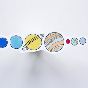 May include: A colorful sticker featuring a hand-drawn illustration of the solar system. The planets are depicted in various colors, including blue, yellow, orange, and red, with black outlines. The sticker is on a white background.