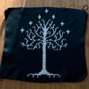 May include: A square dark green crochet blanket featuring a large, centered white tree design with intricate branches and seven small star-like shapes positioned above the canopy, crafted with a consistent stitch pattern throughout the textile.