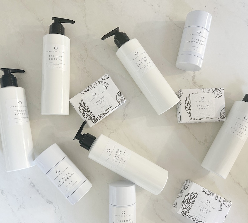 May include: A collection of white bottles and containers with black labels and text that reads "Tallow Lotion", "Tallow Deodorant", and "Tallow Soap". The products are arranged on a white marble surface.