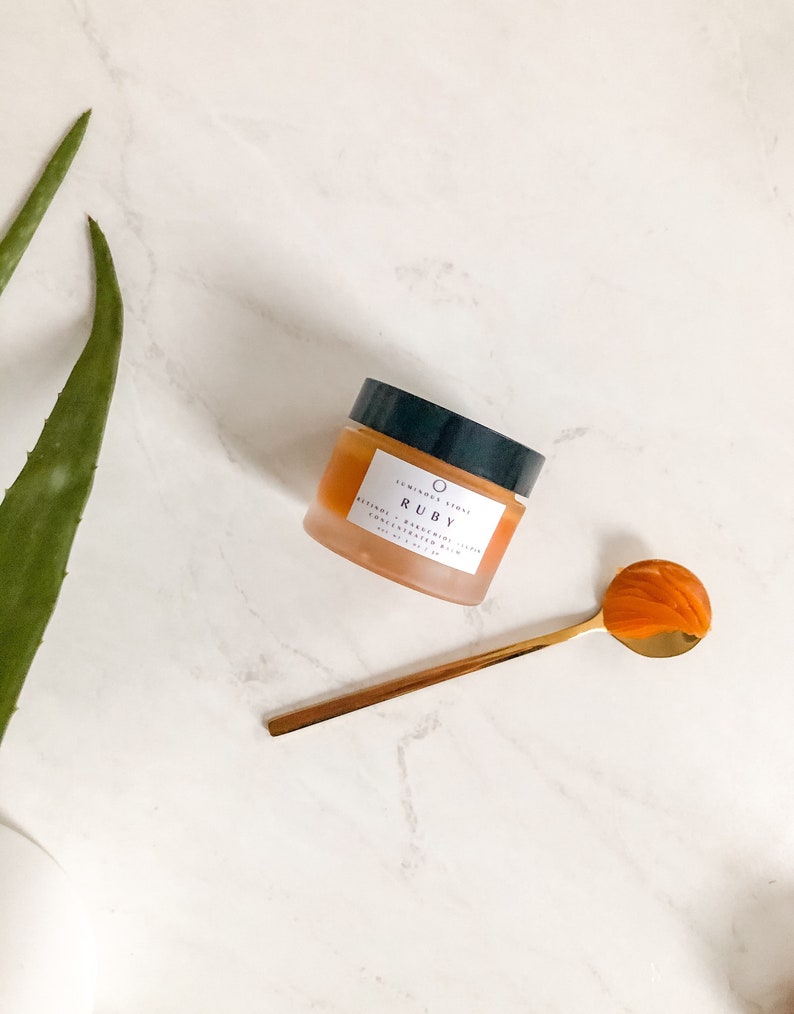 May include: A small jar of orange-colored balm with a black lid and white label that reads "SO MINDS SO SKIN" and "RUBY". The jar is on a white marble surface with a gold spoon beside it.