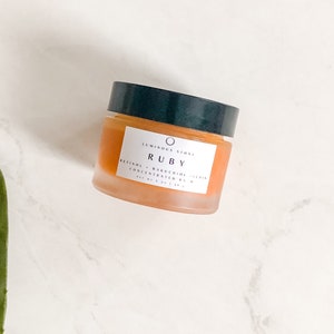 May include: A glass jar of orange-colored skincare product with a black lid. The label reads "Luminous Stone Ruby Retinol + Bakuchiol Complex Concentrated Balm 1 fl oz | 30 ml".