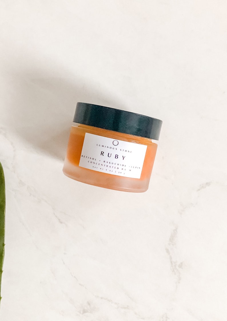 May include: A glass jar of orange-colored skincare product with a black lid. The label reads "Luminous Stone Ruby Retinol + Bakuchiol Complex Concentrated Balm 1 fl oz | 30 ml".