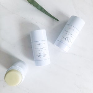 May include: Two white deodorant sticks with a light blue label. The label says "Lemongrass & Patchouli" and "Natural Deodorant". The sticks are on a white marble surface with a green plant leaf in the background.