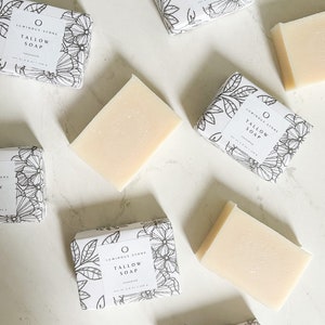Puede incluir: Cinco barras de jab&oacute;n de sebo sin perfume envueltas en papel blanco con dise&ntilde;os florales negros. La etiqueta dice "Luminous Stone Tallow Soap Unscented".
