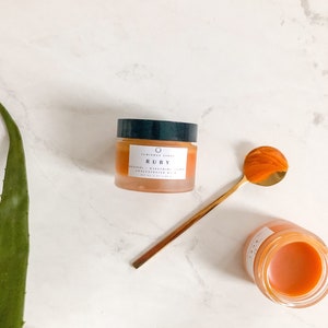 May include: A small jar of orange-colored face cream with the text "LUMINOUS SKIN RUBY RESVERATROL + BAKUCHIOL CONCENTRATE SERUM 20 ml / 0.67 fl oz" on the label. The jar is sitting on a white marble surface with a gold spoon and a green aloe vera plant.