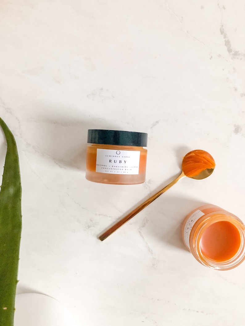 May include: A small jar of orange-colored face cream with the text "LUMINOUS SKIN RUBY RESVERATROL + BAKUCHIOL CONCENTRATE SERUM 20 ml / 0.67 fl oz" on the label. The jar is sitting on a white marble surface with a gold spoon and a green aloe vera plant.