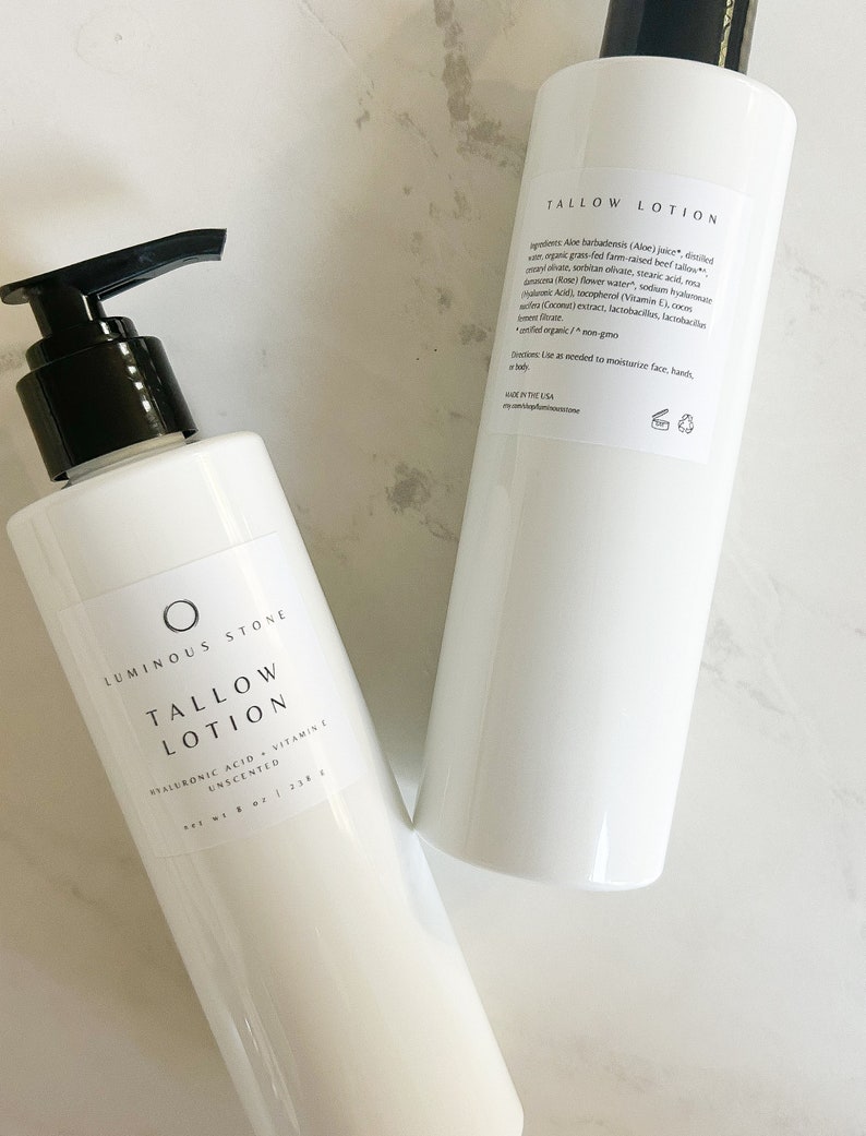 May include: Two white bottles of unscented tallow lotion with black pump tops. The bottle on the right has the ingredients listed on the back. The bottle on the left has the brand name "Luminous Stone" and the text "Tallow Lotion" printed on the front.