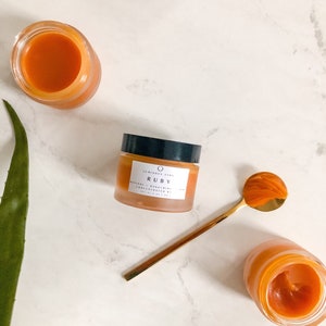 May include: A small glass jar of orange-colored face cream with the label "10 MINDS SKIN RUBY RETINOL & BAKUCHIOL CONCENTRATED SERUM 1.0 FL OZ / 30 ML" on a white marble surface. A gold spoon with a scoop of orange cream is next to the jar.