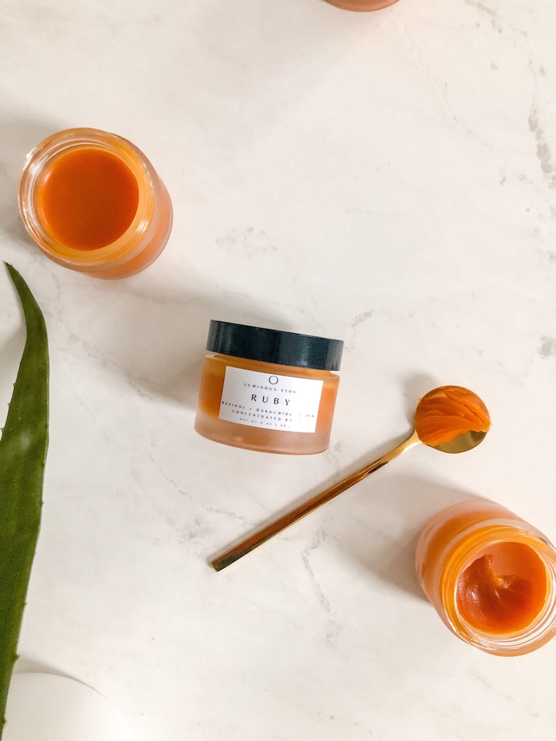 May include: A small glass jar of orange-colored face cream with the label "10 MINDS SKIN RUBY RETINOL & BAKUCHIOL CONCENTRATED SERUM 1.0 FL OZ / 30 ML" on a white marble surface. A gold spoon with a scoop of orange cream is next to the jar.