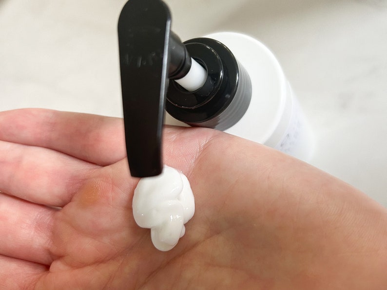 May include: A white pump bottle with a black pump dispenser. A dollop of white lotion is being dispensed onto a hand.