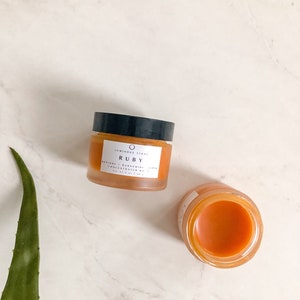 May include: Two glass jars of orange-colored skincare product. The larger jar is labeled "Luminary Stone Ruby Retinol - Bakuchiol Serum Concentrated 1.5%"
