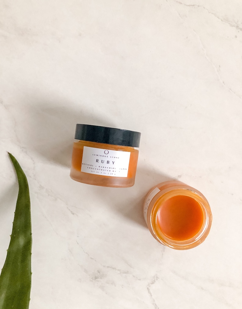 May include: Two glass jars of orange-colored skincare product. The larger jar is labeled "Luminary Stone Ruby Retinol - Bakuchiol Serum Concentrated 1.5%"