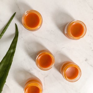 May include: Five small glass jars with orange-colored balm inside. The jars are on a white marble surface.