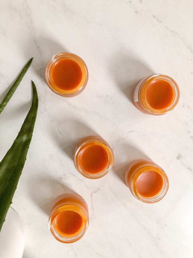 May include: Five small glass jars with orange-colored balm inside. The jars are on a white marble surface.
