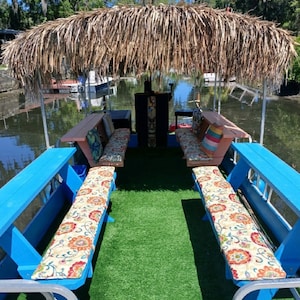 May include: A blue and white boat with a thatched roof and a green artificial grass floor. The boat has two benches with floral patterned cushions.