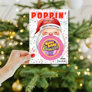 May include: A Christmas-themed card featuring Santa Claus holding a Hubba Bubba Bubble Tape. The card has the word "POPPIN'" in red at the top, with "Merry Christmas!" on Santa's hat. The card is held in front of a Christmas tree.