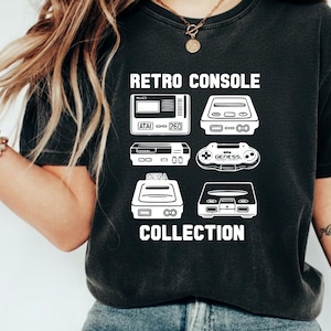 May include: Dark gray t-shirt featuring a white graphic of retro video game consoles. The text "Retro Console Collection" is printed above and below the console illustrations. The shirt is made of a soft material.