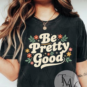 May include: Dark gray t-shirt with the phrase "Be Pretty Good" in a retro font, surrounded by floral and leaf accents in orange, yellow, and green. The shirt has a vintage, worn look.