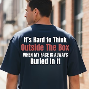 Peut inclure: T-shirt bleu marine avec l'inscription "It's Hard to Think Outside The Box When My Face Is Always Buried In It" en blanc et rouge. Le t-shirt est porté par une personne.