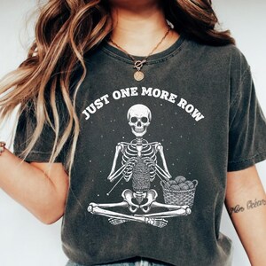 May include: Dark gray t-shirt with a skeleton knitting design. The skeleton is seated cross-legged, holding knitting needles and yarn, with the text "JUST ONE MORE ROW" above. A basket of yarn balls is next to the skeleton.