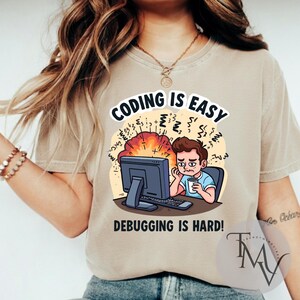 Op de afbeelding: Beige t-shirt met een humoristische print. De tekst "CODING IS EASY" staat boven een illustratie van een gestreste persoon voor een computer. De tekst "DEBUGGING IS HARD!" staat eronder.