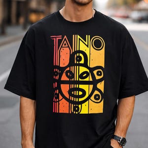 Peut inclure: Un t-shirt noir à manches courtes arborant un imprimé graphique avec le mot TAINO en lettres grasses au-dessus d'un symbole solaire noir stylisé. Le motif est sur un fond dégradé vertical passant de l'orange au jaune avec des lignes texturées.