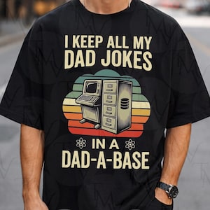 Peut inclure: Un t-shirt noir avec une illustration d'un ordinateur vintage et d'un classeur. Le texte indique I KEEP ALL MY DAD JOKES IN A DAD-A-BASE. Le motif comprend un arrière-plan de style rétro avec des bandes horizontales orange, jaune et vert.
