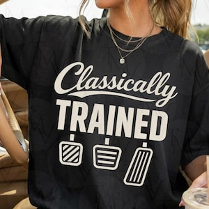 May include: Black t-shirt with the text "Classically TRAINED" in white, above three white pedal graphics. The shirt is a dark color and appears to be made of a soft material. The design is centered on the front of the shirt.
