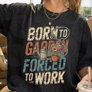 Peut inclure: Un t-shirt noir avec un motif portant le texte BORN TO GARDEN FORCED TO WORK. Le design comprend un arrosoir métallique et un pot en terre cuite rempli de fleurs colorées et de feuilles vertes, reliés par un tuyau d'arrosage vert.