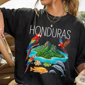 May include: A black t-shirt featuring the white text HONDURAS above a graphic illustration of a tropical island, two red and blue macaws, a striped angelfish, a yellow fish, and two gray dolphins swimming in blue water.