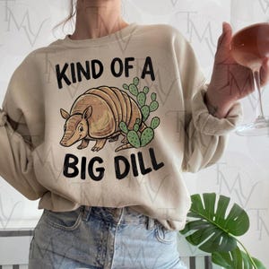 May include: Beige sweatshirt with the text "KIND OF A BIG DILL" and a cartoon armadillo illustration with green cacti. The armadillo is brown and tan. The sweatshirt is a neutral color and has long sleeves.