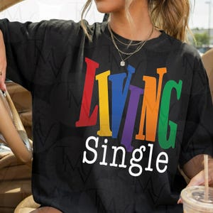 May include: Black t-shirt with the words "LIVING" in rainbow colors and the word "Single" in white. The shirt has a relaxed fit and short sleeves. The design is centered on the front of the shirt.