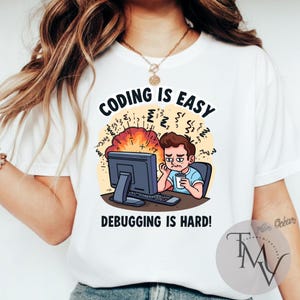 Op de afbeelding: Wit T-shirt met de tekst "CODING IS EASY, DEBUGGING IS HARD!" met een cartoon van een gestreste persoon voor een computer. Het ontwerp bevat een computer, toetsenbord en een kopje koffie.