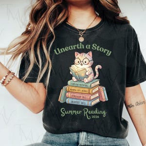 May include: Dark gray t-shirt featuring a cartoon cat wearing glasses and reading a book atop a stack of books. The text "Unearth a Story" and "Summer Reading 2026" are printed above and below the cat illustration. The books have titles like "Storyland Adventures".