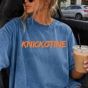 Peut inclure: Un t-shirt bleu à manches courtes avec un aspect délavé et texturé. L'avant du vêtement présente le mot KNICKOTINE imprimé en lettres orange vif et grasses sur la poitrine. Le t-shirt est doté d'une encolure ronde classique.