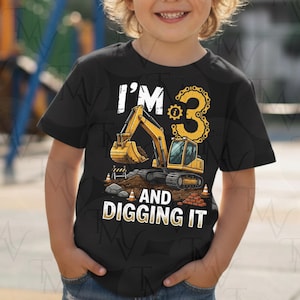 Puede incluir: Una camiseta negra de manga corta con un gráfico de excavadora amarilla, conos de tráfico naranjas y el texto "I'M 3 AND DIGGING IT" con el número tres estilizado como una cadena de engranajes amarilla, sobre tierra y ladrillos.