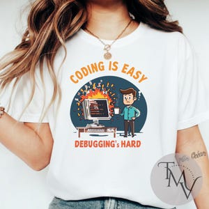 Op de afbeelding: Wit t-shirt met een afbeelding van een brandende computer, een persoon met een mok en de tekst "CODING IS EASY DEBUGGING'S HARD" in oranje en wit. Het ontwerp is geplaatst tegen een blauwe cirkel.
