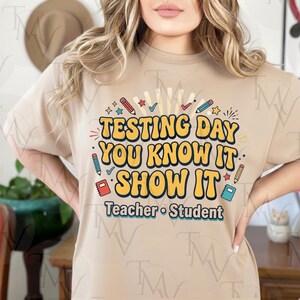 Peut inclure: Un t-shirt beige avec le texte "TESTING DAY YOU KNOW IT SHOW IT" en jaune et bleu, avec des graphiques sur le thème de l'école. Les mots "Teacher • Student" sont en dessous du texte principal.