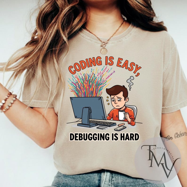 Op de afbeelding: Beige t-shirt met de tekst "CODING IS EASY, DEBUGGING IS HARD." De afbeelding toont een gestreste cartoonpersoon achter een computer met kleurrijke code die uit het scherm komt. De persoon houdt een mok vast en er komt stoom uit zijn hoofd.